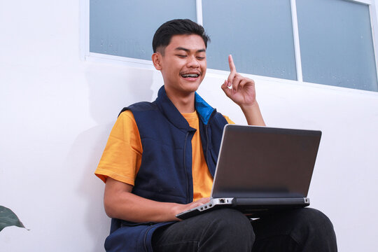 Cheerful young Asian student male with braces teeth and modern laptop on knee pointing up showing got an idea gesture while sitting outdoor