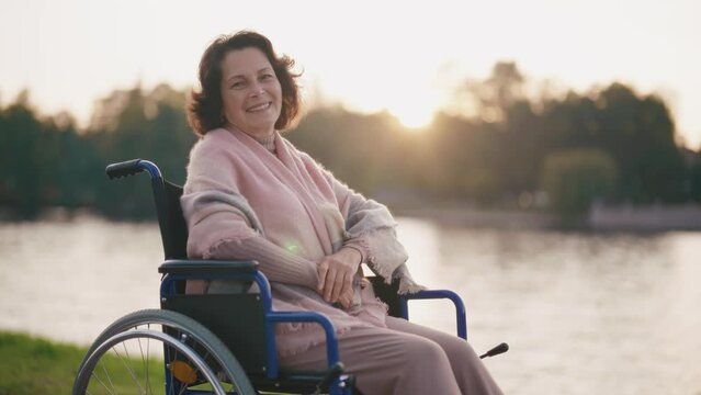 Senior woman in wheelchair smiling on nature. Happy elderly mature grandmother enjoy a walk at sunset look camera. Happiness old, living with legs paralysis, special needs pensioner care concept.