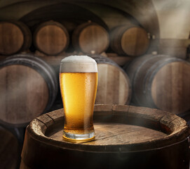 Glass of beer on top of  wooden barrel and  wood barrels in cellar at the background. Sun ray falling down to it surface. Placement for your product.