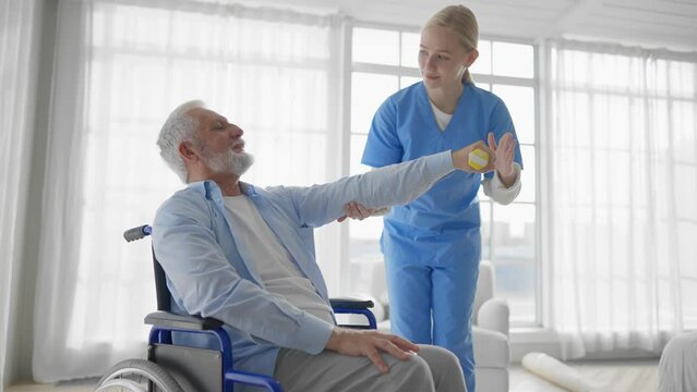 Nurse helps old man in a wheelchair perform physical therapy exercises. A female rehabilitation specialist provides medical assistance to paralyzed man. Palliative care of neurodegenerative pathology.