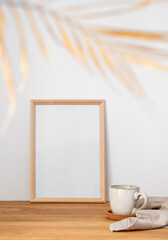 A frame with a blank canvas against a light wall and on a wooden tabletop with blurred palm leaf and cup of tea.