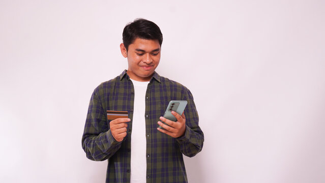 Adult Asian man smiling while holding blank credit card and mobile phone over gray background - Powered by Adobe