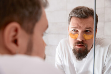 Portrait of man in white tshirt applying eye patches on his face at bathroom. Self care morning...