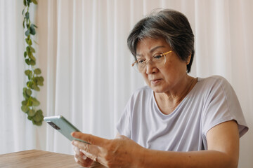 Asian Thai Chinese elder woman using mobile phone while sitting on chair, wear eyeglasses reading news from cellphone.