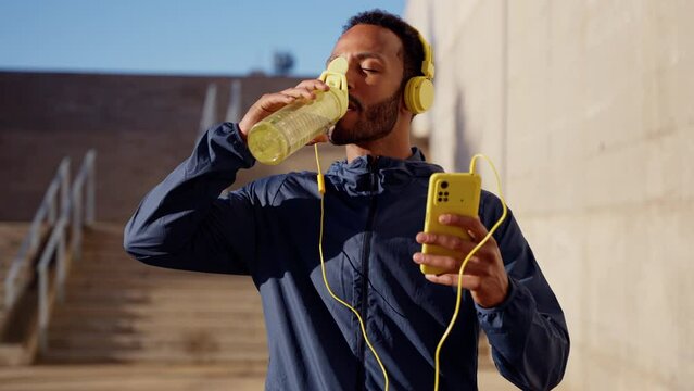 Athletic man with headphones tired from exercising takes a break to drink water while using a cell phone app.