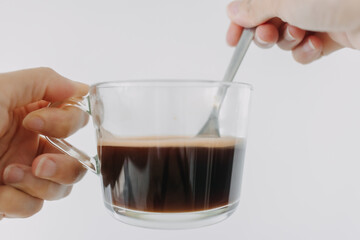 Close up of woman hand holding hot americano coffee clear mug with tea spoon, drink isolated over white background wall.