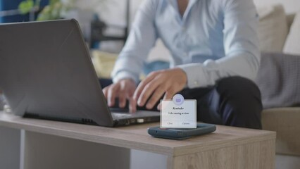 Mobile phone Video meeting reminder popup distracting Young adult freelancer business man working from home in his living room using laptop, wearing a light blue Button-Up Shirt