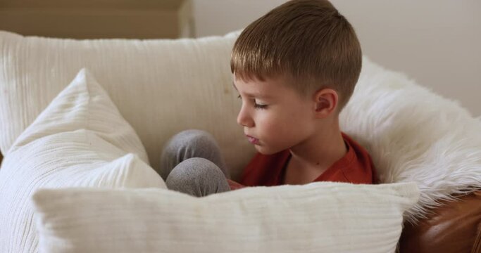 Close up shot little 7s sad offended Caucasian boy sitting on armchair surrounded with cushions feels frustrated, not in mood to play or talk, looks disgruntled and upset. Childhood grievances, whims