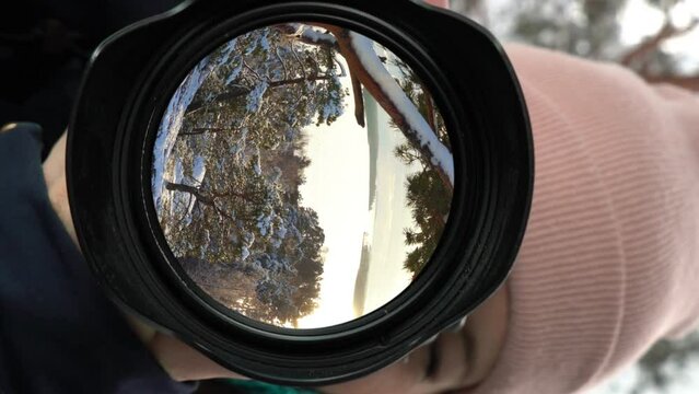 A young woman photographer takes a picture in a winter forest, snow-covered pines and sunset are reflected in the lens as in a mirror.