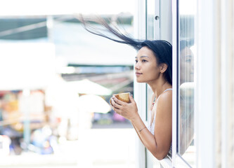 A happy woman in a summer dress holds a mug in an open window of a residential building while the wind blows her hair