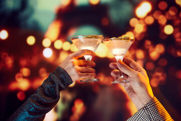 Romantic date. Mae and female hands clinking martini cocktails over bokeh background. Cheers. Concept of holidays, celebration, events, Christmas, New Year, Valentines Day