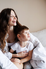 Portrait of smiling woman hugging cute toddler son under blanket on bed in morning