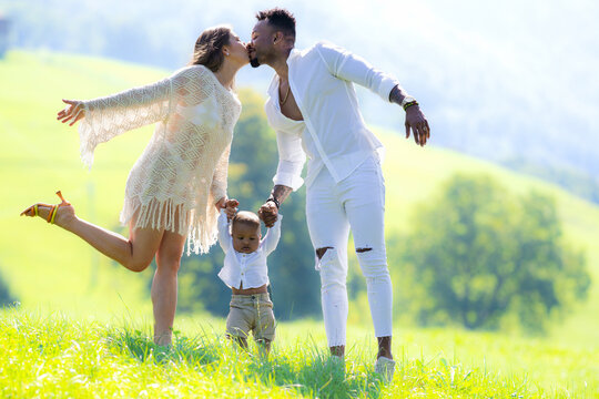 Multiethnic parents and mixed race baby walking on summer grass. Multiethnic couple kissing on nature. Biracial Multiethnic baby child first step on backyard. Diverse Multiethnic family.