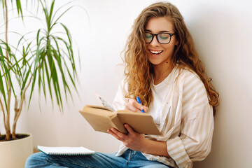 Young woman reading a book, writing notes, doing homework at home.  Business, blogging, freelance, education concept. Concept of rest, relaxation, comfort. - obrazy, fototapety, plakaty