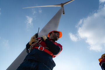 Low angle view of a male engineer standing in front of a wind turbine, Using the walkie talkie to...