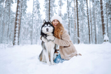 A young woman in warm clothes walking her dog in a picturesque snowy forest. Woman laughing and playing with pet in the park. Domestic dog concept.