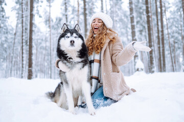 A young woman in warm clothes walking her dog in a picturesque snowy forest. Woman laughing and playing with pet in the park. Domestic dog concept.