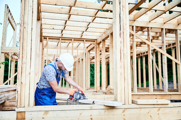 Fototapeta premium Carpenter using circular saw for cutting wooden plank. Man worker building wooden frame house. The idea behind modern ecological construction.