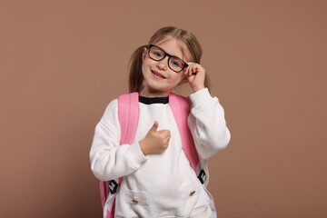 Happy schoolgirl in glasses with backpack showing thumb up gesture on brown background