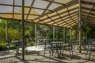 A sunshade-covered decking area with dining chairs and tables. Outdoor verandah eating area of a casual restaurant in the countryside with a view of Australian bushland or woods.