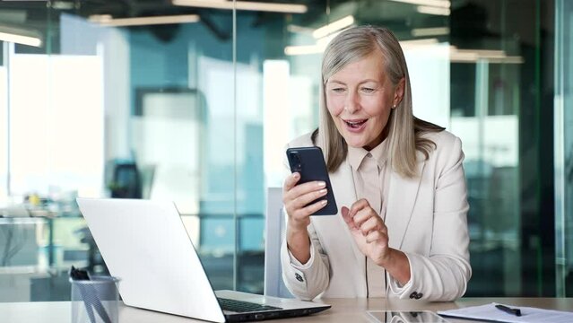 Happy excited senior gray haired businesswoman read great news on smartphone sitting at workplace in business office. Mature business female celebrates success, rejoices at the win, yes, triumph, wow