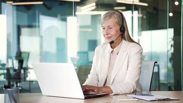 Senior gray haired female corporate operator who works in support service speaks on a video chat using a laptop. A call center agent in a wireless headset helps a customer with a complaint in office