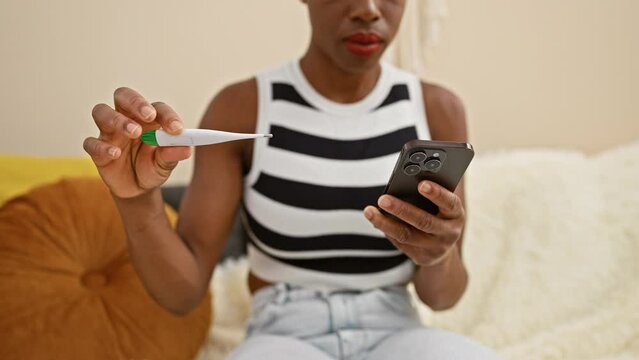 Concerned African American Woman At Home, Taking Temperature With A Thermometer, Glued To Her Smartphone Screen - Teleconsulting, Messaging Amid Fever