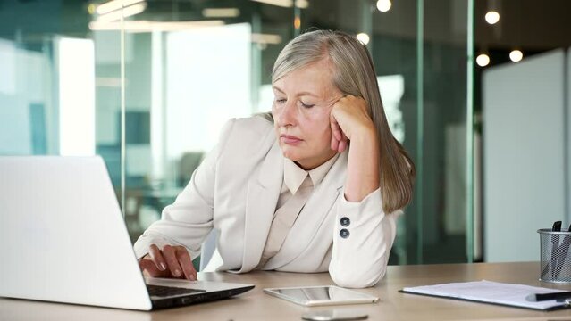 Overworked elderly senior gray haired businesswoman is bored sitting at workplace at desk in business office. Sleepy exhausted woman is tired from working with laptop, procrastinates, gets distracted