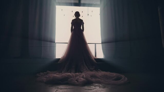 mysterious woman in purple luxurious dress long train walks in dark room towards window balcony. Fantasy girl princess back rear view evening gown go to light. Royal queen silhouette bride wedding. 4k