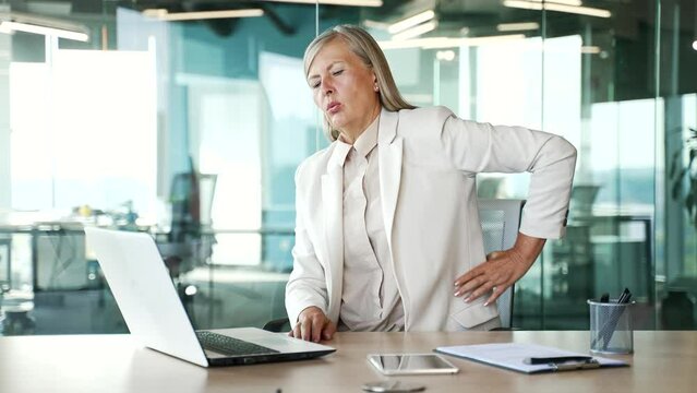 Tired elderly senior gray haired businesswoman suffering from back pain working on laptop sitting at workplace in office. Upset mature female holds her hand on her lower back, massaging sore muscles
