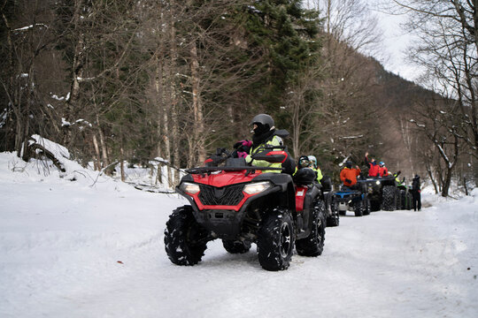 A man, equipped with cold-weather attire, confidently handles a red quad bike along a snowy path, surrounded by a wintry forest landscape.