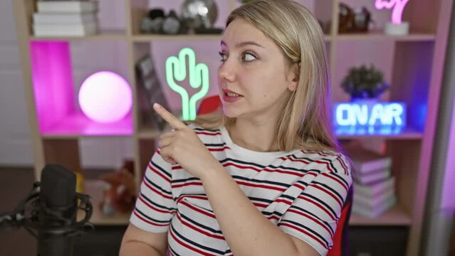 Shocked young blonde woman streamer gesturing nervously, pointing aside at computer screen with worried expression - chaos in gaming room!