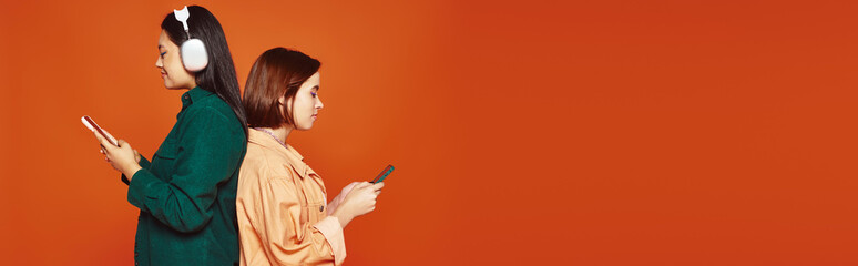 happy asian girl in wireless headphones holding smartphone near her female friend on orange, banner