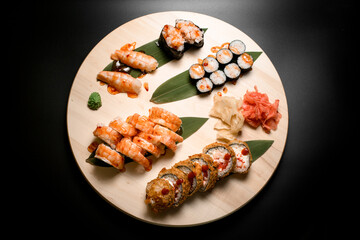 Top view of a set of sushi rolls on a round wooden board with wasabi, red and white ginger