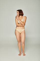 full length of shy woman in beige underwear looking over shoulder on grey background, vulnerability