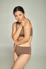 young asian woman in underwear covering body while embracing herself on grey backdrop, body shaming