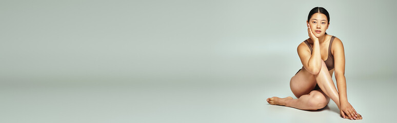 Obraz premium full length of young asian woman in underwear sitting with crossed legs on grey background, banner