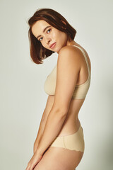 charming young woman with short hair standing in beige underwear and looking at camera on grey