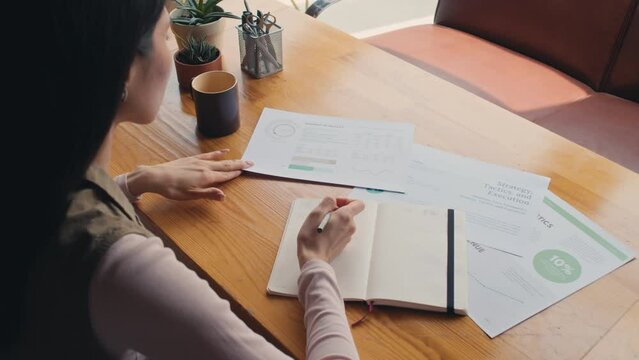 Medium over-shoulder shot of young Caucasian woman sitting alone at table, studying presentation sheets with financial tables and graphs, and writing notes in workbook