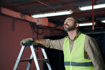 handsome pensive technician in safety vest standing next to step ladder and looking up, data center