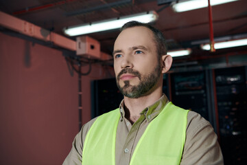 attractive pensive specialist in safety vest posing and looking away while working in data center