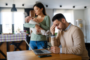 Father businessman try to work on laptop from home with wife and baby on background.