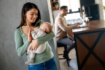 Young father works on laptop while his wife looks after an infant child. Work from home