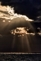 Fototapeta premium The sun shines through dark clouds creating beams of light in the air and on the water.