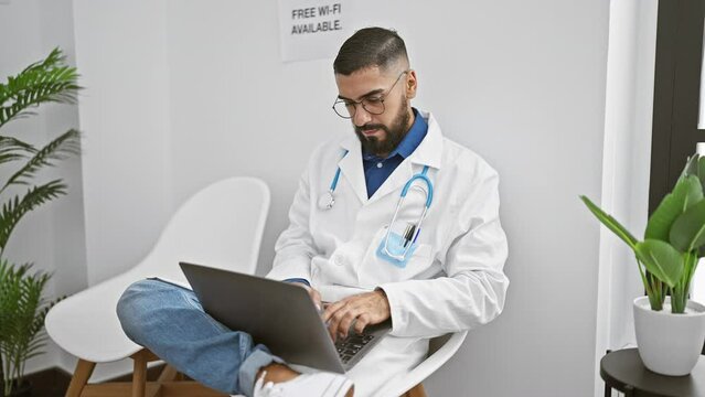 A bearded man in a white lab coat works on a laptop in a modern clinic with plants and free wi-fi sign visible.