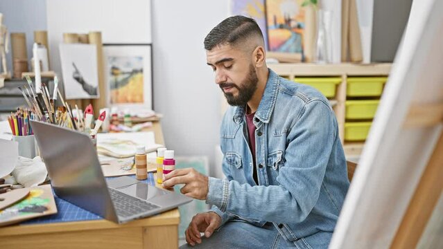 Bearded man examines paint in art studio with laptop and easel.