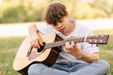 Handsome teen boy is learning how to play guitar while sitting in park on grass.