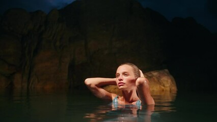 Ethereal beauty in blue dress immersed in night sea, rocks behind. Elegant lady enjoys serene sea bath under moonlight. Skin care, luxury fashion scene. Mystic coastal ambiance, tranquil water moment.