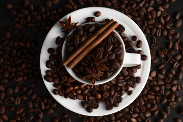 On a black background in a coffee cup there are coffee grains, cinnamon sticks and cardamom fruits.
