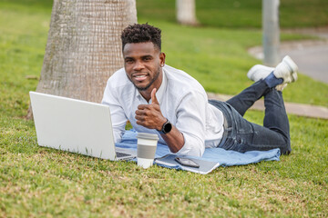 You see an African-American man lying on the green grass smiling and thumbs up, engrossed in his work on a laptop.